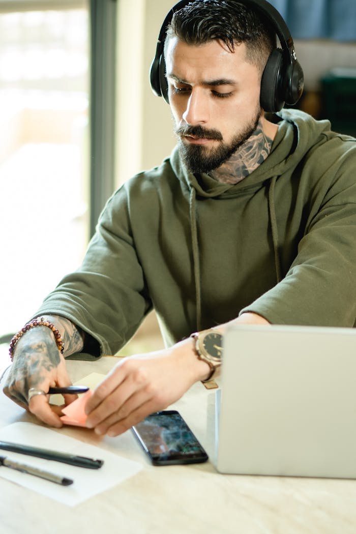 Bearded man with tattoos and headphones working on a laptop in a home office setting.
